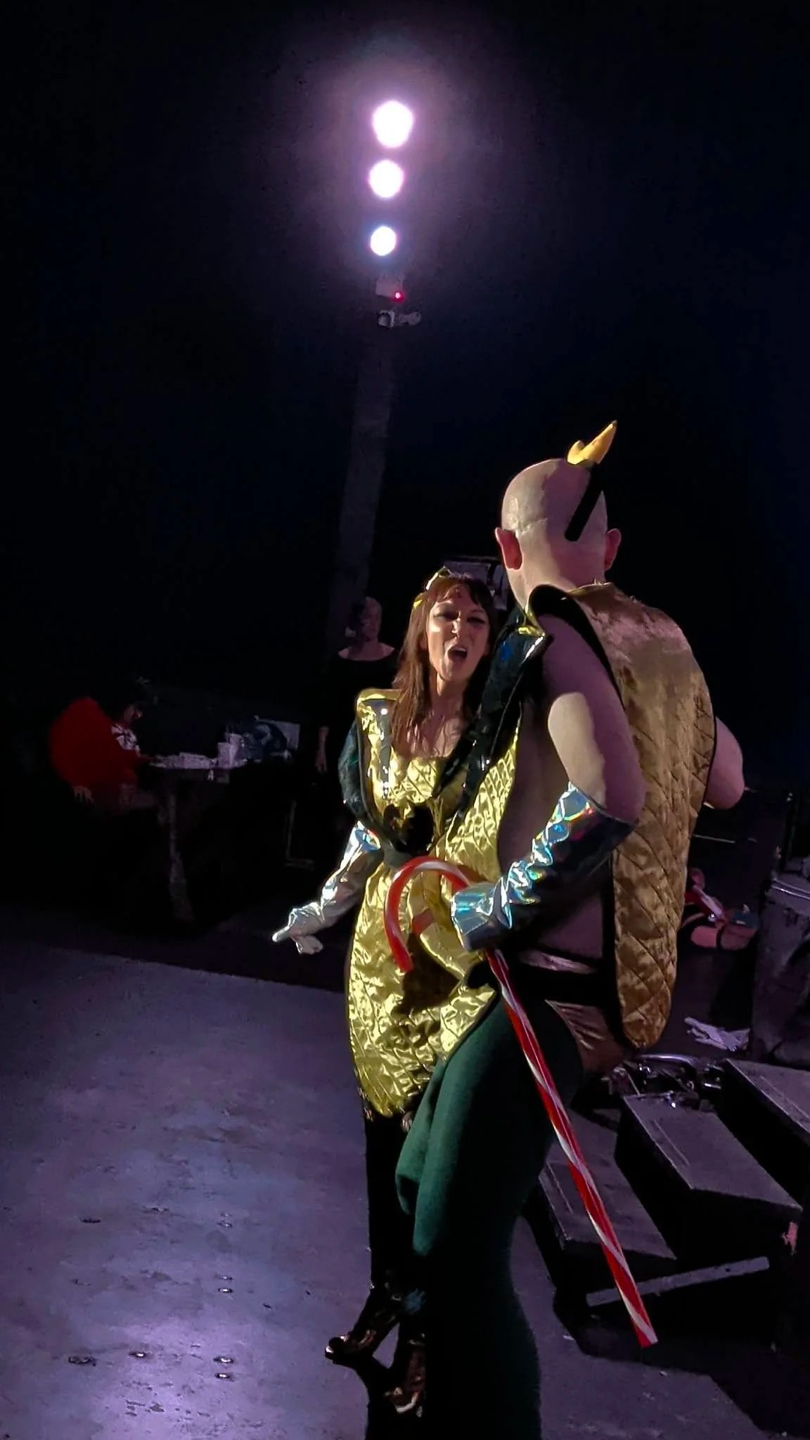 Stage lighting illuminates the scene. A man in gold and silver costuming dressed as Magenta from the Rocky Horror Picture Show faces a man dressed in gold and silver costuming dressed as Riff Raff. They are performing.