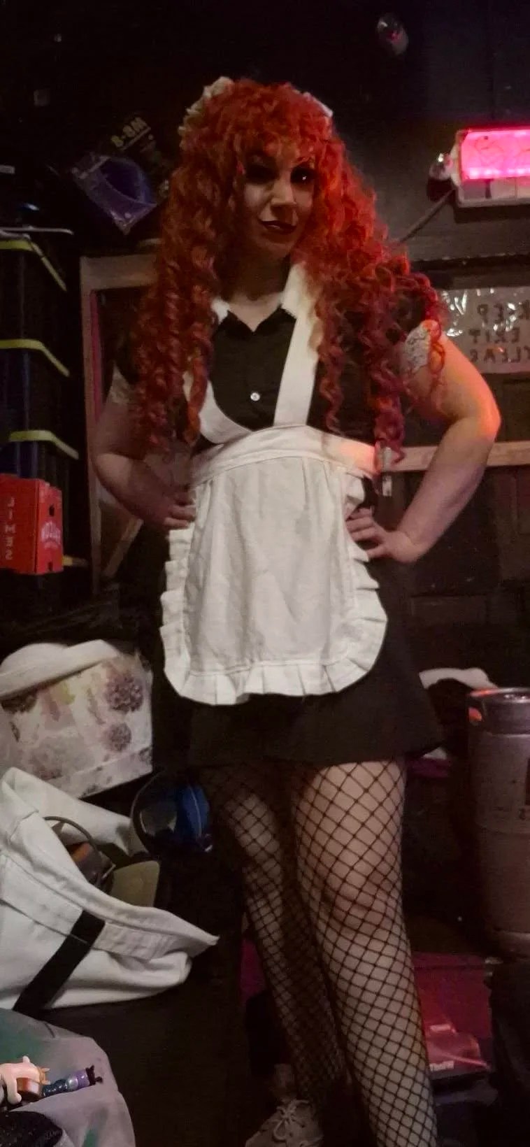 Person dressed as the maid Magenta from The Rocky Horror Picture Show. They are wearing a red, curly wig and a black dress with a white apron, and fishnet stockings. They have both hands on their hips.